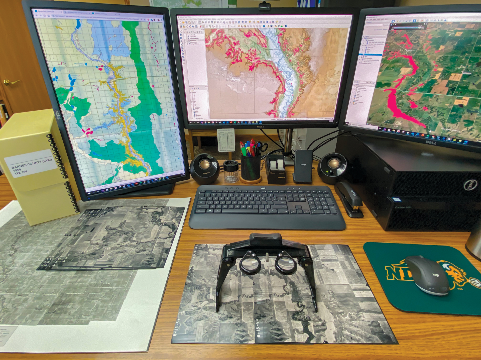 Desk with black and white aerial photos and three computer screens displaying digital, colored maps.