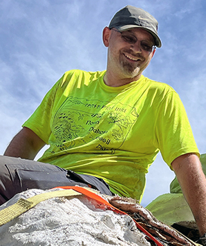 Photo of Clint Boyd sitting on a plaster fossil jacket with blue sky in the background.
