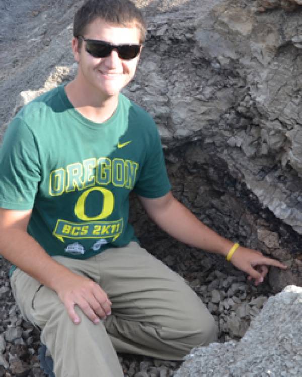 Sean Ternes wearing a green t-shirt and khaki pants pointing to a fossil in the dirt.