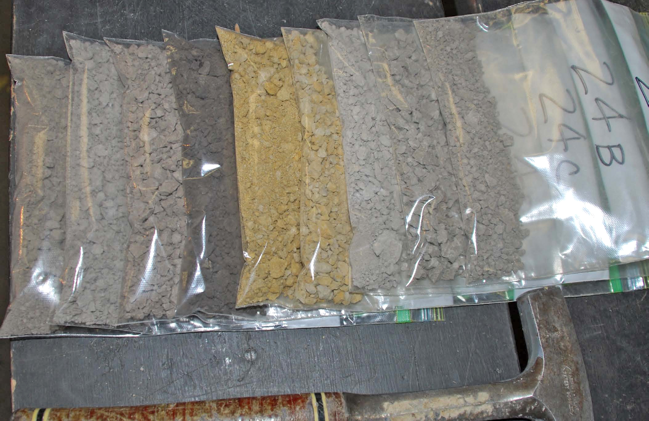 Nine clear plastic bags filled with grayish brown, dark brown and yellowish rocks on a table next to a rock hammer. 